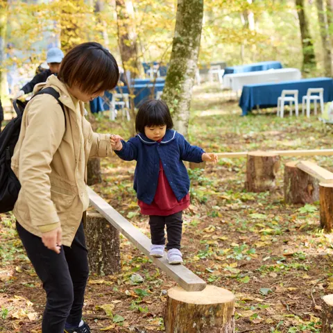 森の中の木の平均台を、小さな子が大人に手を引かれて渡っている。