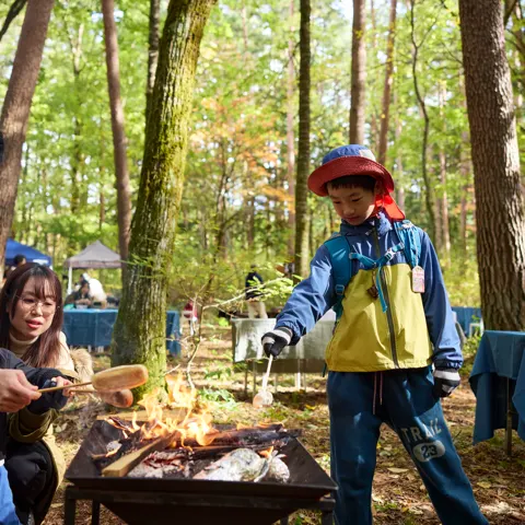 森の中のたき火台で、大人と子どもが串に刺したソーセージを焼いている。