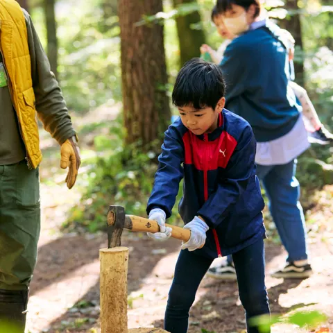森の中で、男の子が斧を構えて台の上の薪を割ろうとし、横で大人が見守っている。