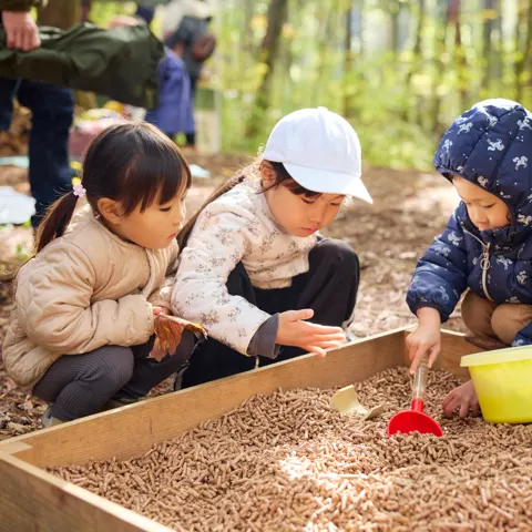 木の枠のペレット砂場で、3人の子が黄色いバケツやスコップを使って遊んでいる。