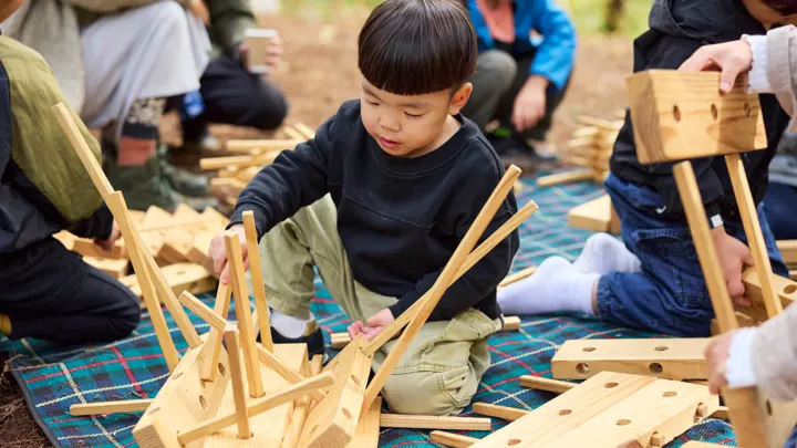 青いシートの上で、男の子が木の積み木やおもちゃを組み立てている。