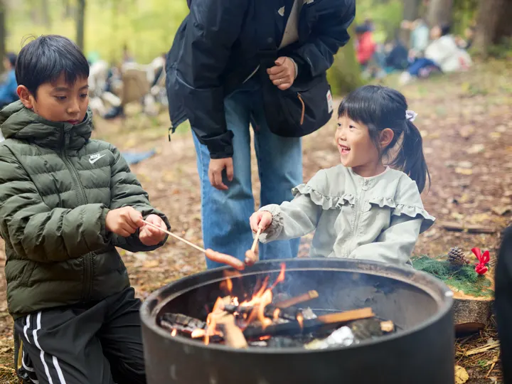 黒いドラム缶のたき火で、男の子と女の子が串を火にかざして焼いている。