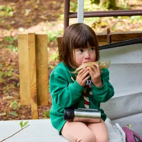緑のトレーナーを着た幼い子が、紙に包んだサンドイッチを両手で持って食べている。