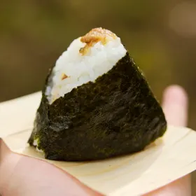 手のひらにのせた木目のある薄い板の上に、のりを巻いた三角のおむすびが置かれ、てっぺんに茶色い具がのっている。