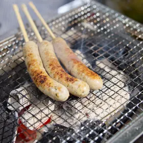 炭火の網の上で、串に刺した手作りソーセージが3本焼けている。