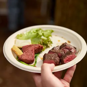 丸い紙皿に、焼いた鹿肉とレタスなどの野菜が盛られ、手で支えて持っている。