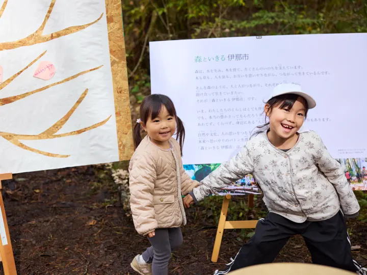 木の絵ボードの前で、白い帽子の女の子が笑って立ち、隣にもう1人の子がいる。