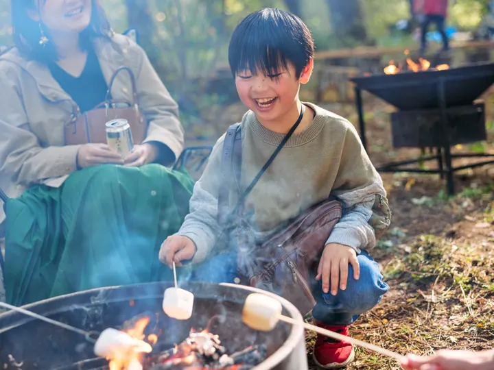 たき火台の前で、男の子が串に刺したマシュマロを焼きながら笑っている。
