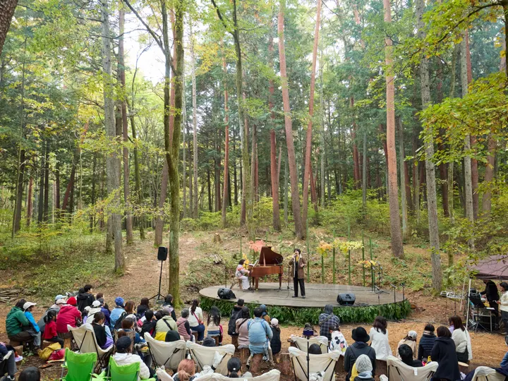林の中の円形ステージでピアノと歌の演奏があり、客席の人が座って聴いている。