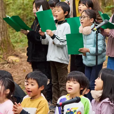森の中で、子どもたちがマイクの前に並び、口を大きく開けて歌っている。